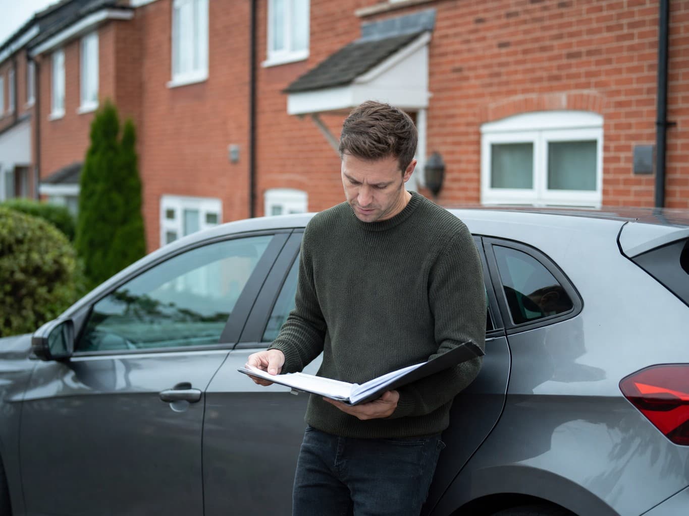 A car owner standing on a UK driveway examining paperwork beside their car