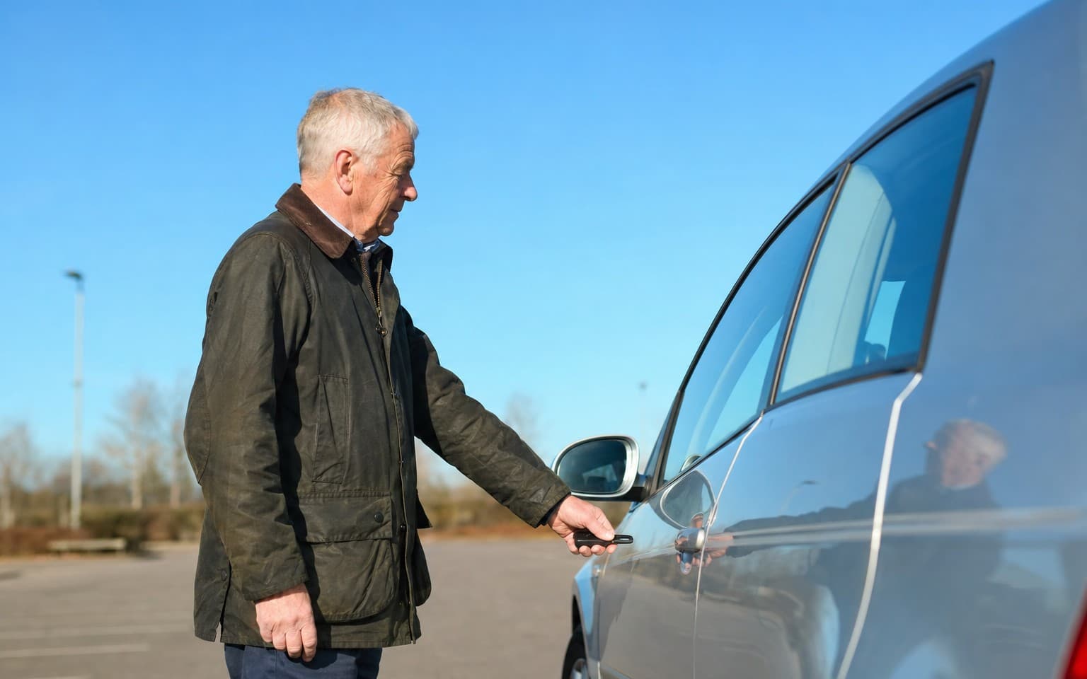 An older man opening the door of a grey saloon in an empty car park to look inside