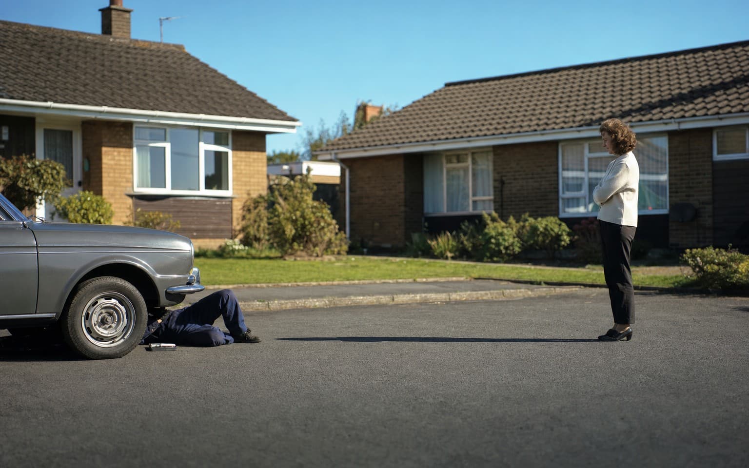An inspector on his back under the front of a grey saloon in a cul-de-sac while the owner watches from the drive
