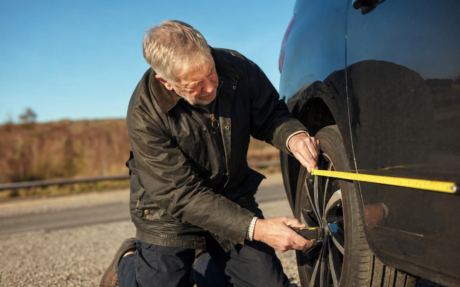 A man in a waxed jacket kneeling at the wheel of a black MPV in a lay-by, measuring the tyre with a tape