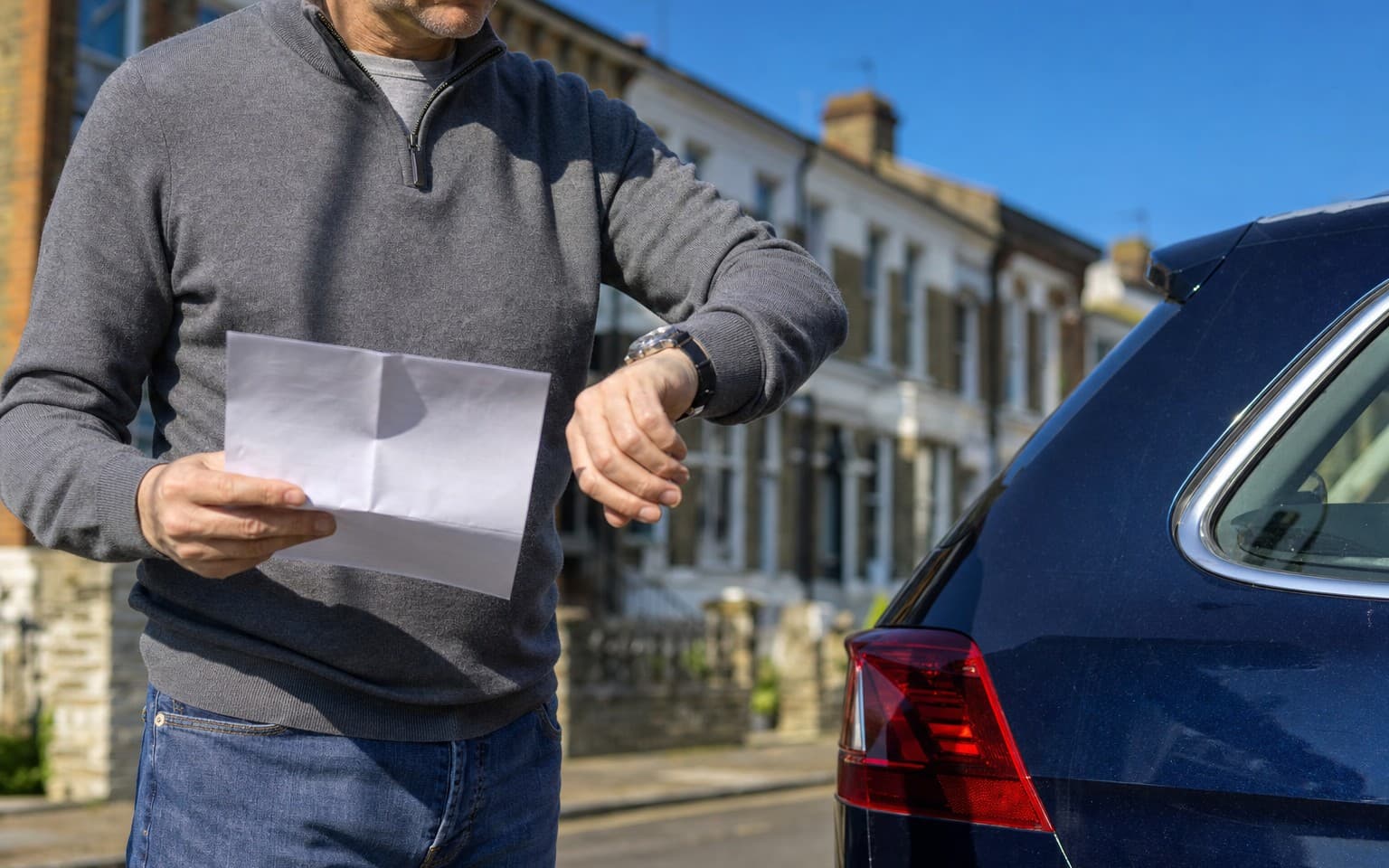 A man checking his watch with a letter in his other hand beside his dark blue estate on a street of terraces