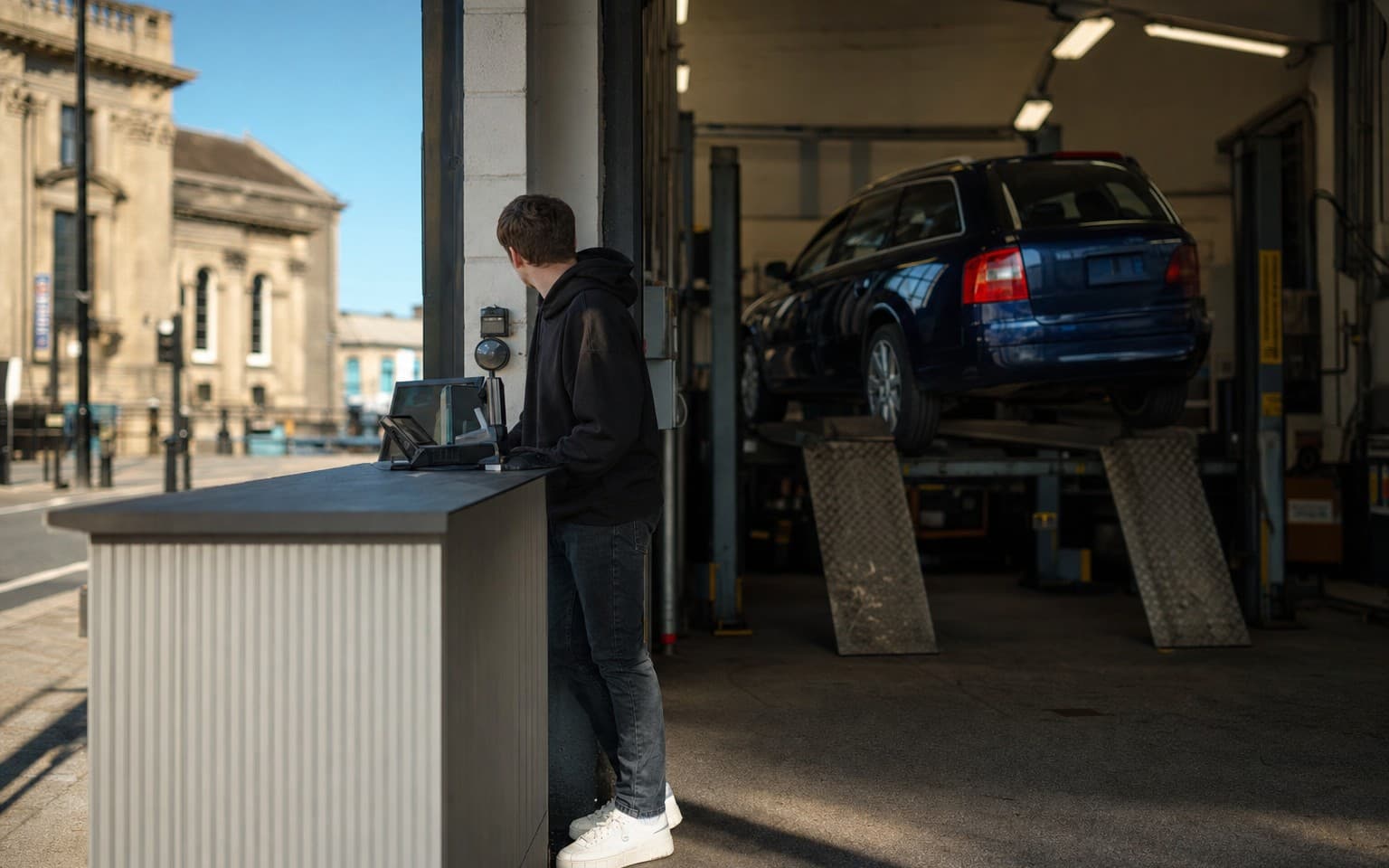A young man at the counter of a garage while his dark blue estate sits on the lift behind him